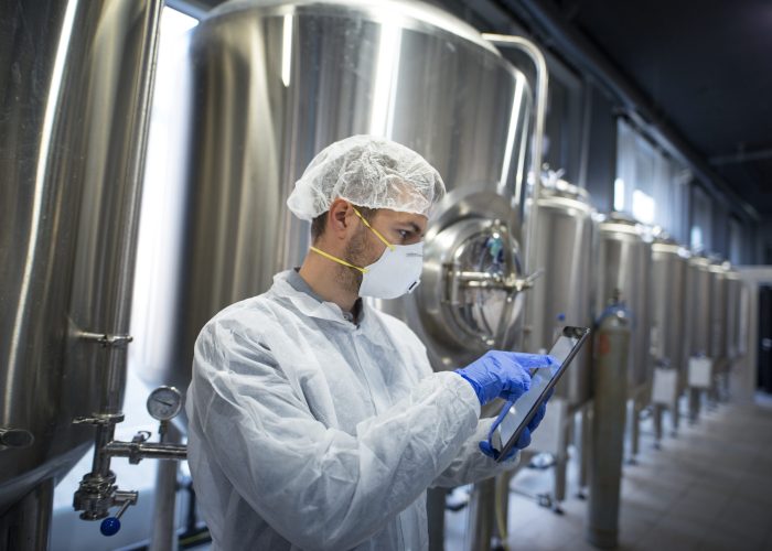 Technologist in protective suit holding tablet and checking production in factory plant.